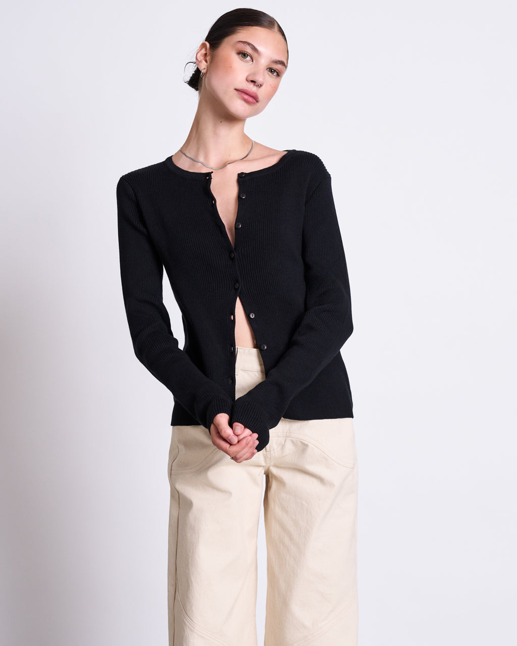 A person stands against a plain light background wearing the jan-n-june CREWNECK CARDIGAN GLIMT BLACK GOTS with top buttons undone and cream wide-leg pants, exuding comfort in organic cotton as they clasp their hands and tilt their head slightly.