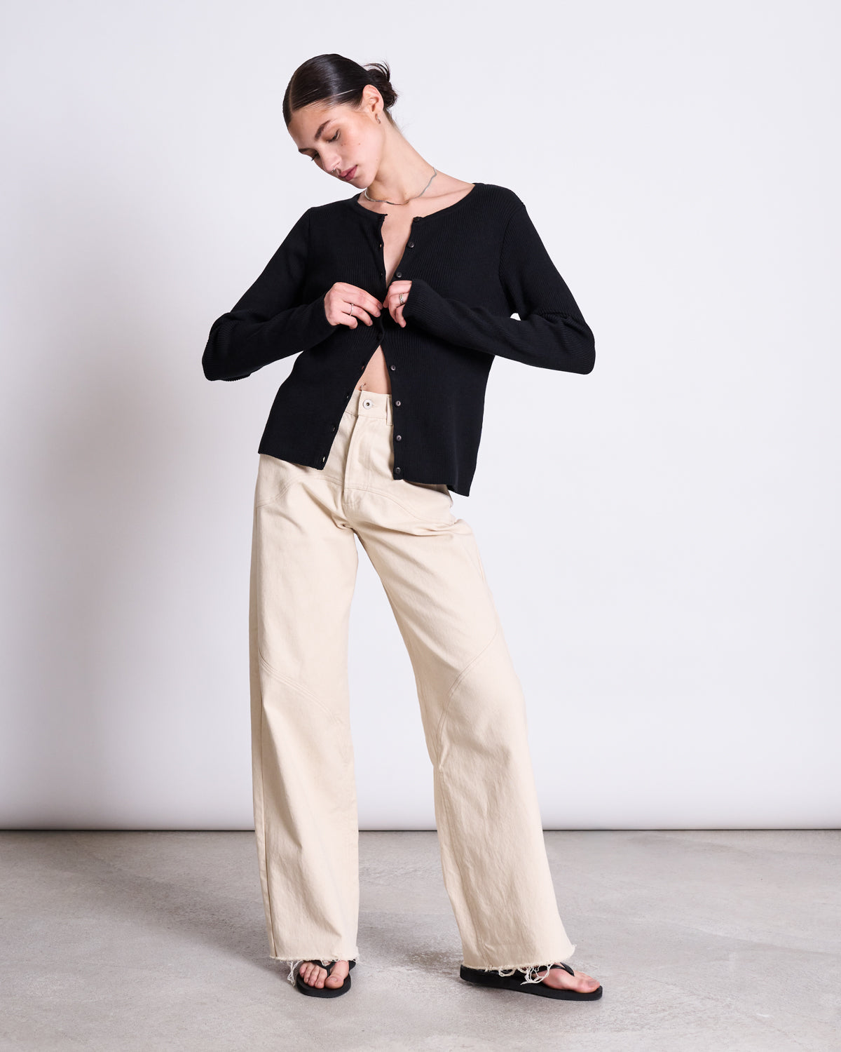 A woman stands indoors against a plain background, wearing the jan-n-june CREWNECK CARDIGAN GLIMT BLACK GOTS, beige wide-leg pants, and black sandals. She is looking down while casually buttoning her cardigan.