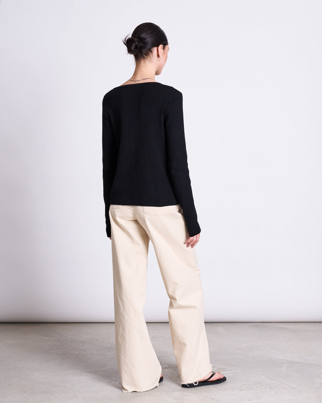 A person with dark hair in a bun stands facing away, wearing the jan-n-june CREWNECK CARDIGAN GLIMT BLACK GOTS, beige wide-leg pants, and black sandals in a minimalist indoor setting with a light background.