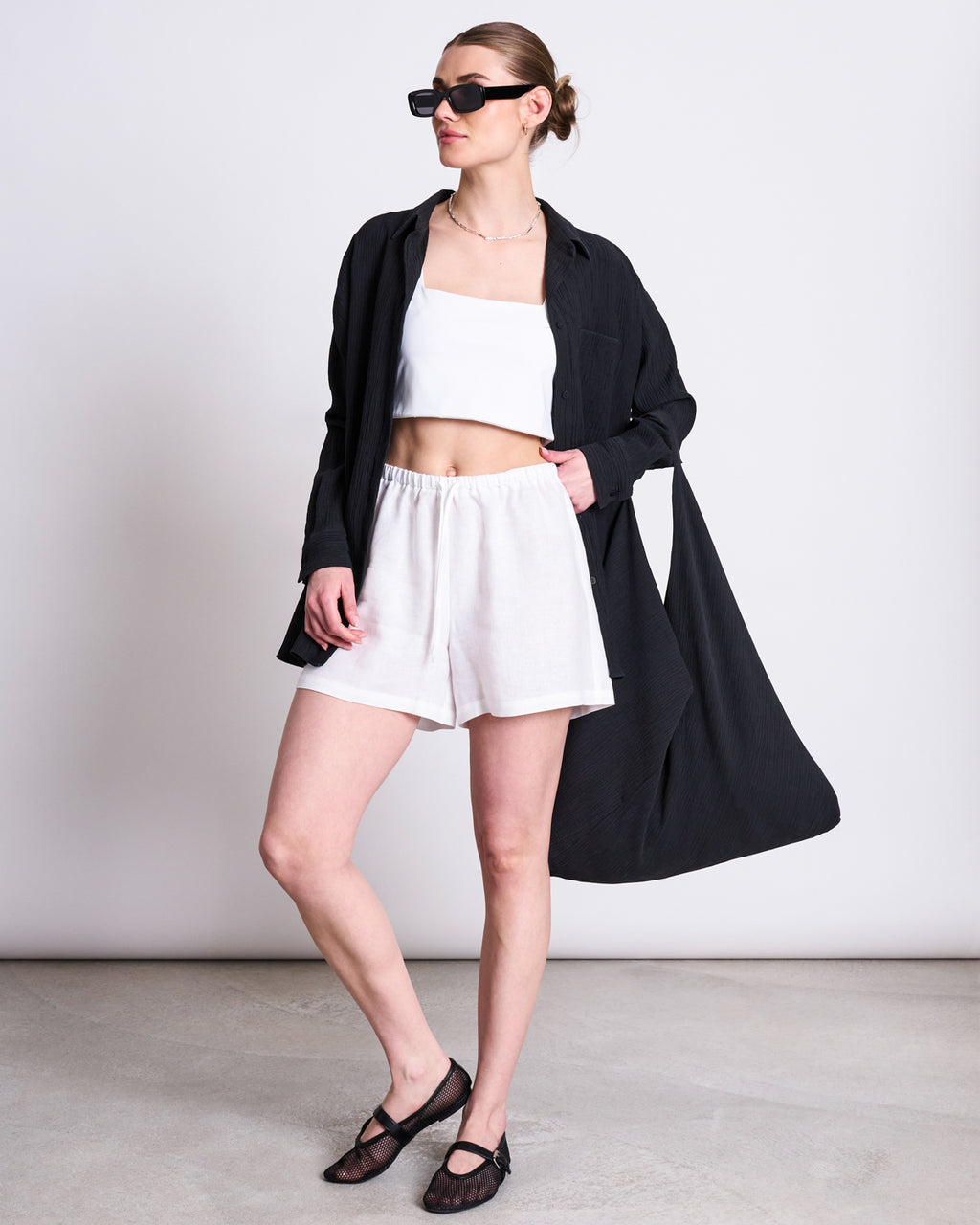 A woman poses against a plain background in black sunglasses, the jan-n-june CRINKLE SHIRT SKUR BLACK, a white crop top, white shorts, and black flats. Her hair is pulled back and she has one hand in her pocket.