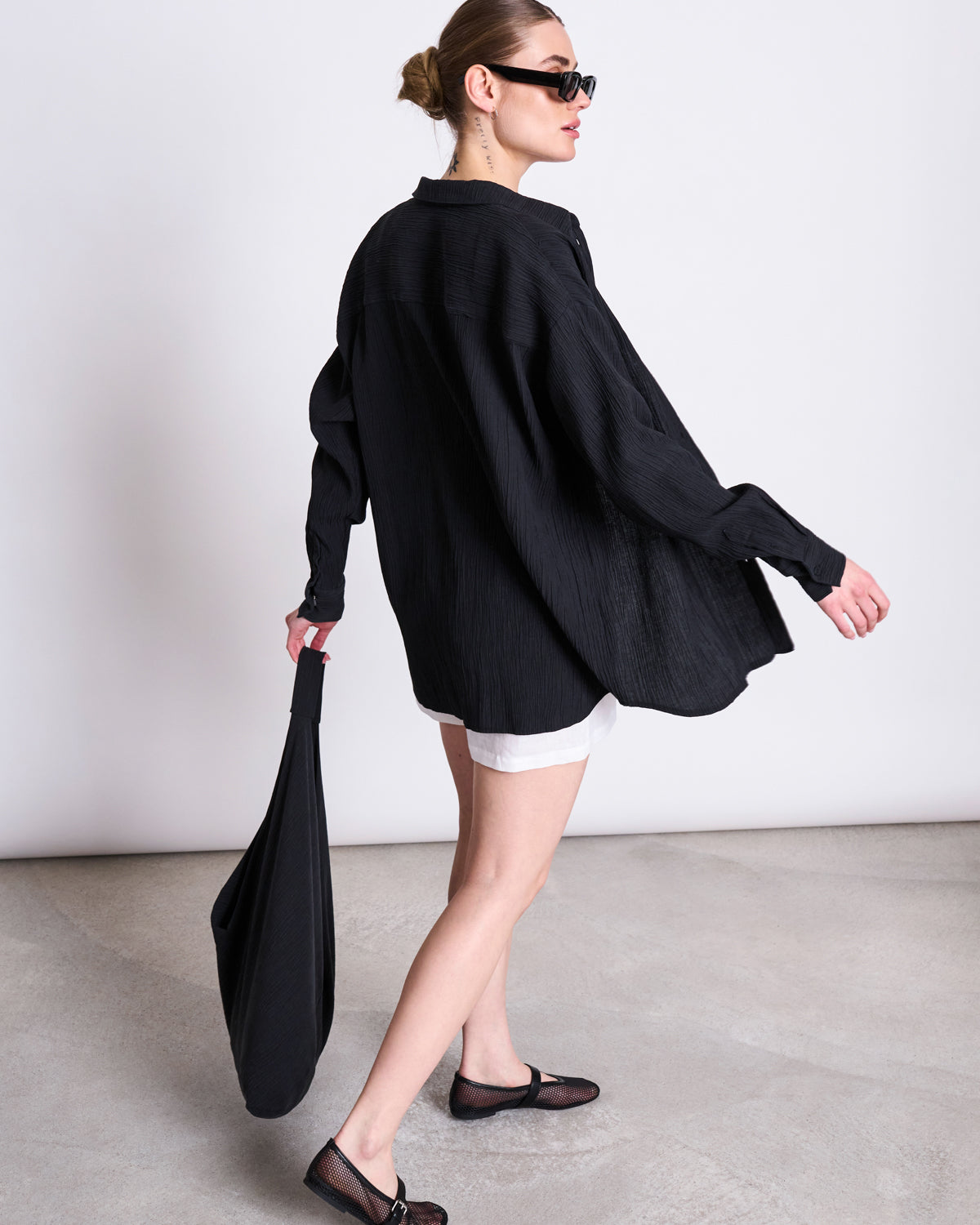A person in sunglasses, a jan-n-june CRINKLE SHIRT SKUR BLACK, white shorts, and black shoes walks on a light floor, carrying a black bag at their side.