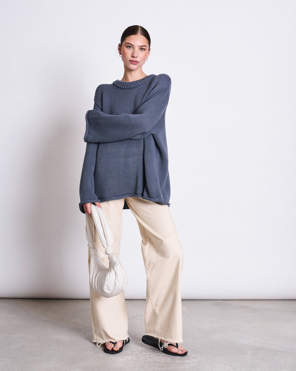A woman stands against a plain white wall wearing the jan-n-june JUMPER SONA TURBULENCE GOTS, paired with wide-leg beige pants and black sandals. She holds a white puffy handbag and looks confidently at the camera.