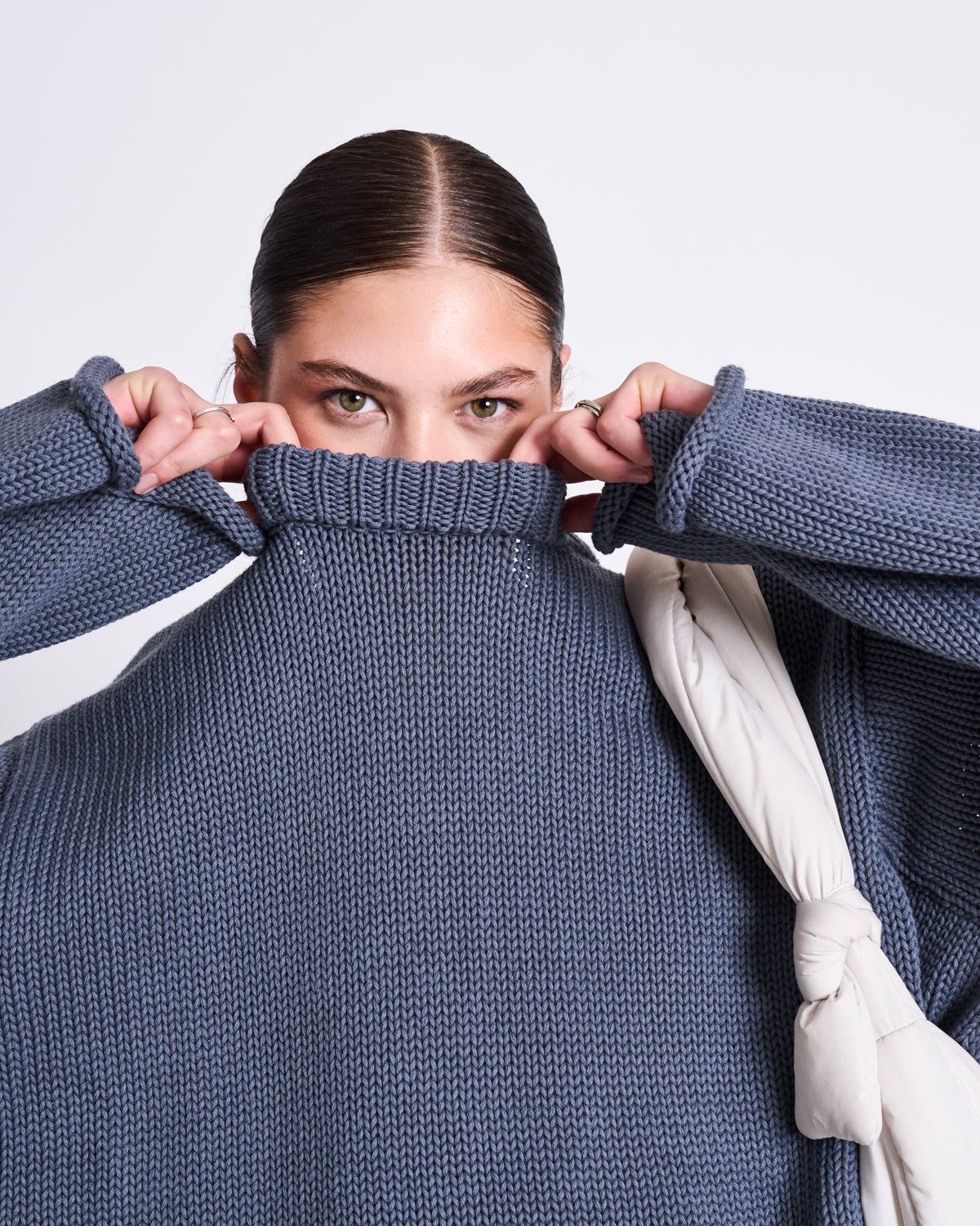 A person with slicked-back hair pulls up the high neck of the oversized JUMPER SONA TURBULENCE GOTS by jan-n-june, partially covering their face. They have intense green eyes and carry a white, knotted bag on their shoulder.