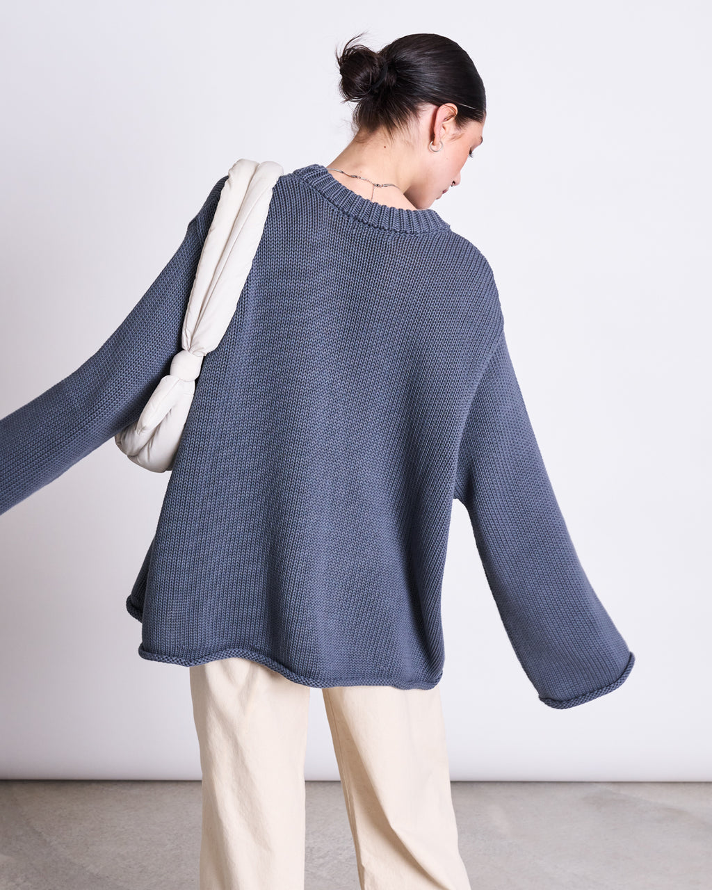 A person with dark hair in a low bun wears the jan-n-june JUMPER SONA TURBULENCE GOTS, an oversized blue-gray knitwear piece, paired with light beige pants and a white padded shoulder bag, standing against a plain light background.
