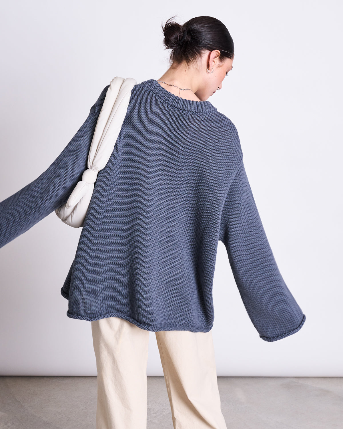 A person with dark hair in a low bun wears the jan-n-june JUMPER SONA TURBULENCE GOTS, an oversized blue-gray knitwear piece, paired with light beige pants and a white padded shoulder bag, standing against a plain light background.