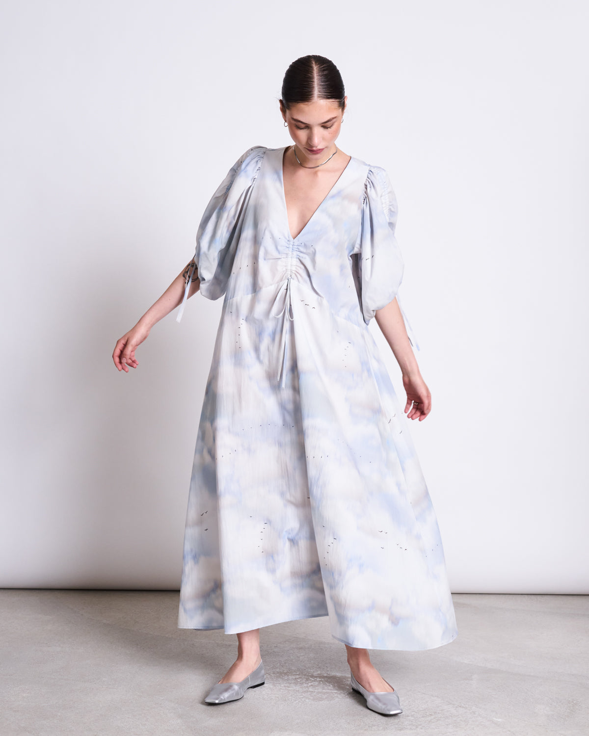 A woman in the jan-n-june MAXI DRESS DANS CLOUD PRINT—a pale blue and white maxi with puffed sleeves and a deep V-neck—stands against a white background, gazing down and holding her dress while wearing gray flats.