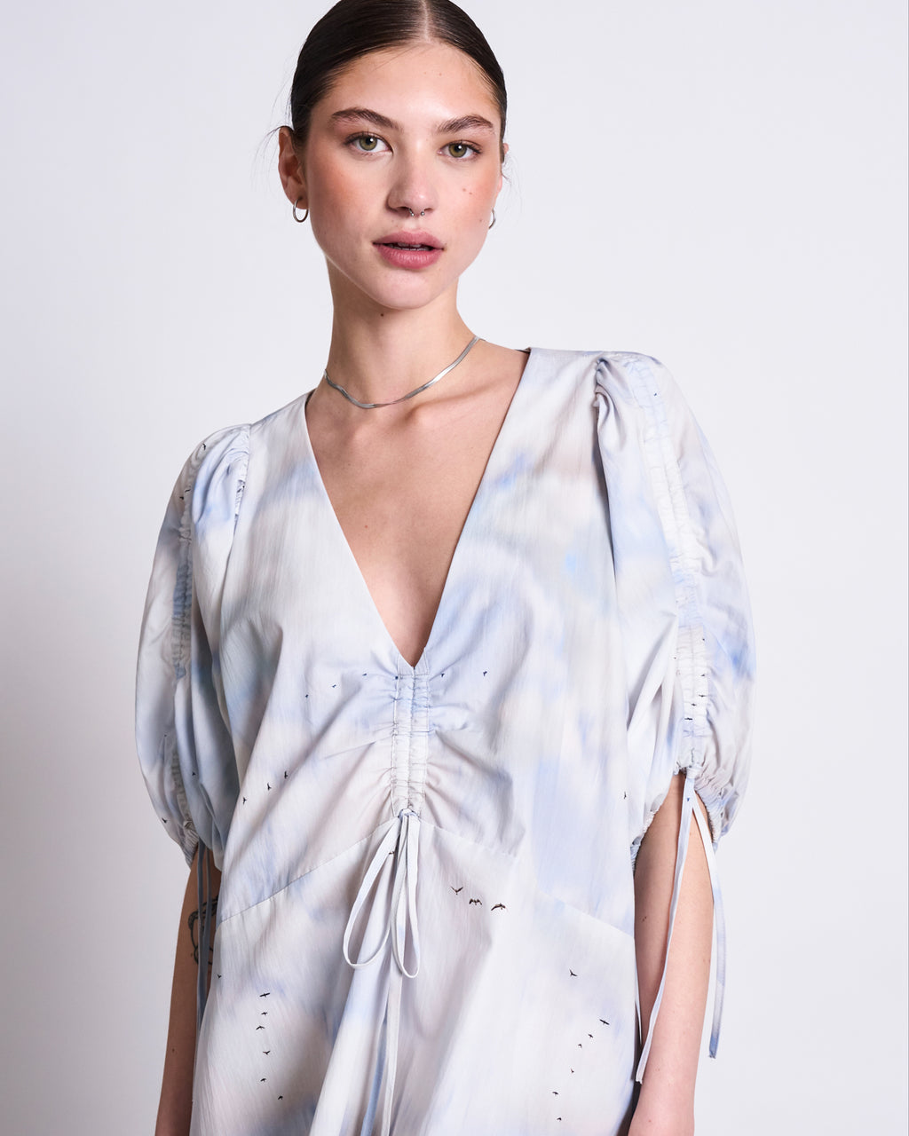 A woman with straight brown hair tied back wears the jan-n-june MAXI DRESS DANS CLOUD PRINT, a pale blue and white organic cotton v-neck maxi dress with ruched sleeves and drawstring detail, posing against a plain light background.