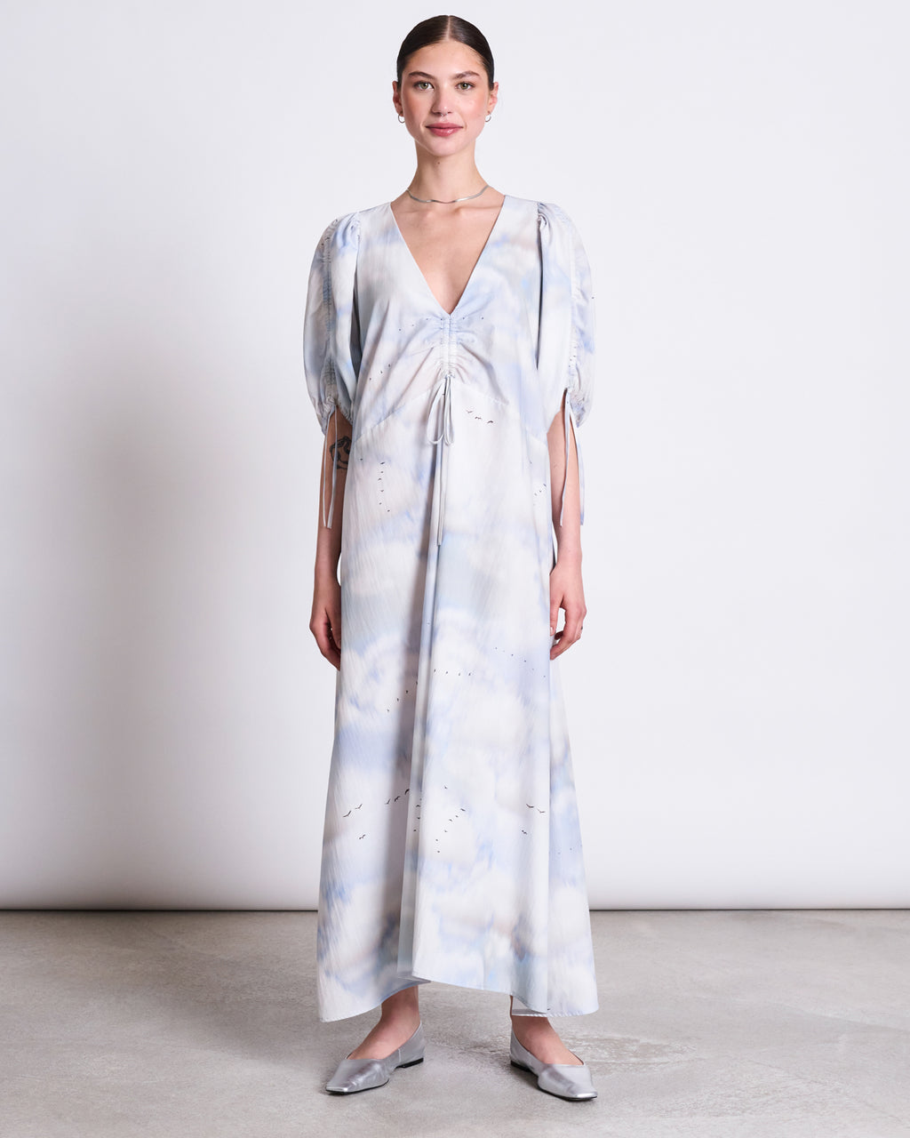 A woman faces forward and smiles slightly, wearing jan-n-junes MAXI DRESS DANS CLOUD PRINT—an organic cotton maxi with a light blue and white cloud pattern, deep V-neck, puffed half sleeves—and silver flat shoes against a plain background.