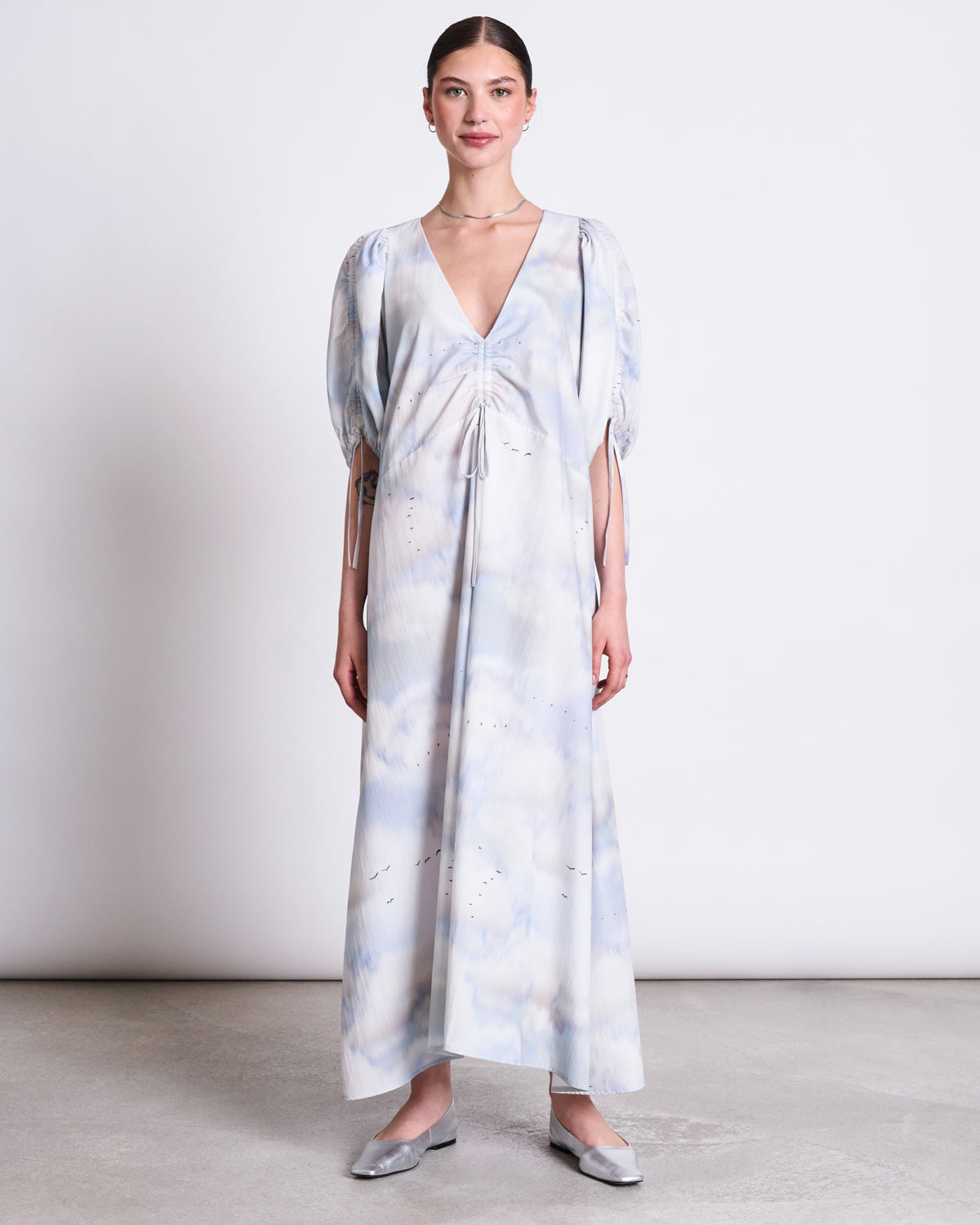 A woman faces forward and smiles slightly, wearing jan-n-junes MAXI DRESS DANS CLOUD PRINT—an organic cotton maxi with a light blue and white cloud pattern, deep V-neck, puffed half sleeves—and silver flat shoes against a plain background.