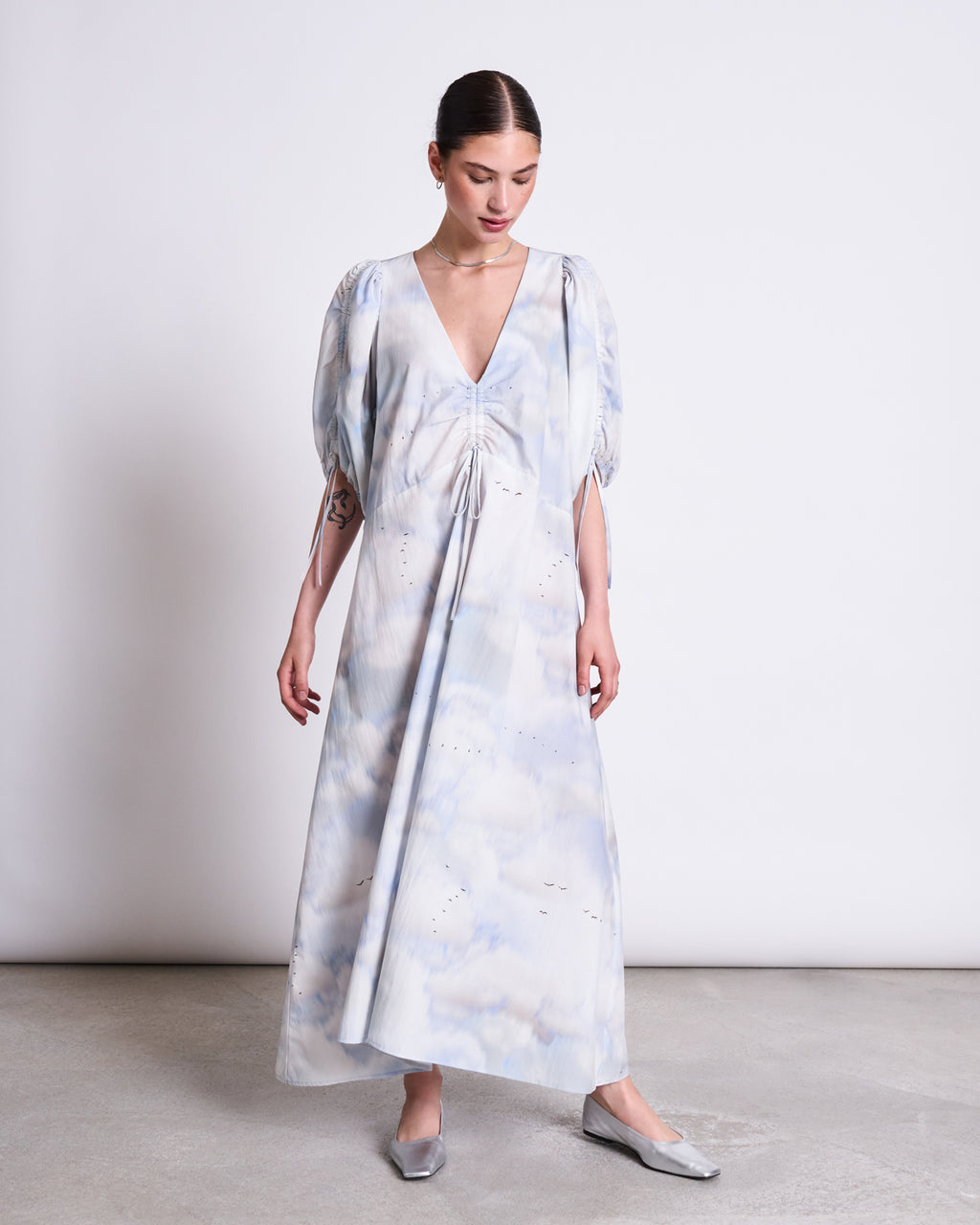 A woman stands indoors wearing the jan-n-june MAXI DRESS DANS CLOUD PRINT, a flowing organic cotton maxi with light blue and white cloud pattern, short puff sleeves, deep V-neck, and silver flats. The flattering length complements her silhouette.