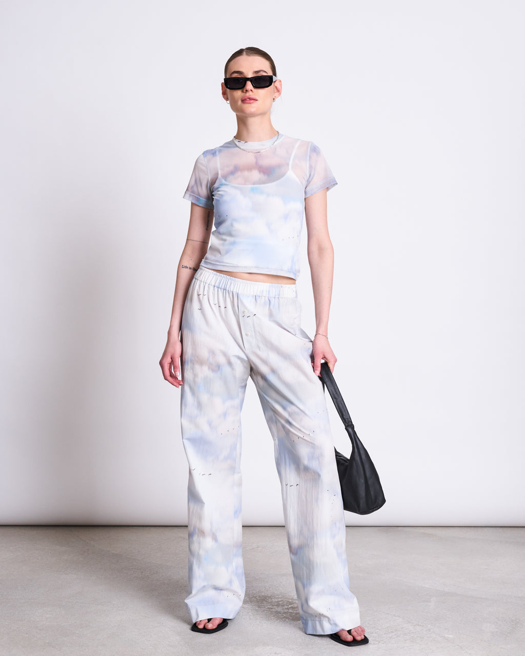 A woman in the jan-n-june MESH T-SHIRT EVIE CLOUD PRINT, layered over a white bralette with matching tie-dye pants, black sandals, sunglasses, and a black purse stands against a white wall—perfect festival wear.