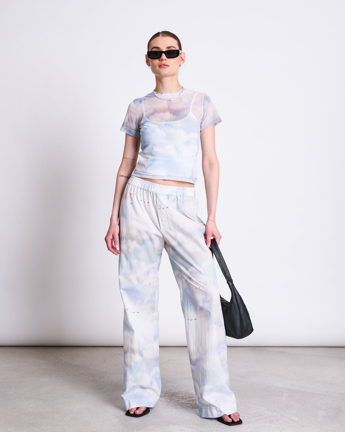 A woman in the jan-n-june MESH T-SHIRT EVIE CLOUD PRINT, layered over a white bralette with matching tie-dye pants, black sandals, sunglasses, and a black purse stands against a white wall—perfect festival wear.