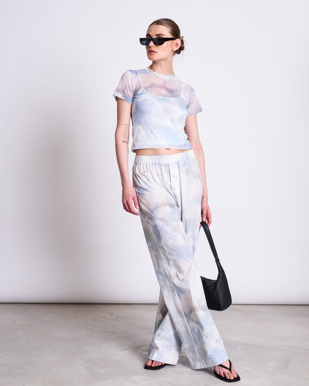 A woman wears the jan-n-june MESH T-SHIRT EVIE CLOUD PRINT with matching wide-leg pants. Perfect for festivals, she pairs her recycled mesh outfit with sunglasses, black sandals, and a black bag, standing confidently against a plain background.