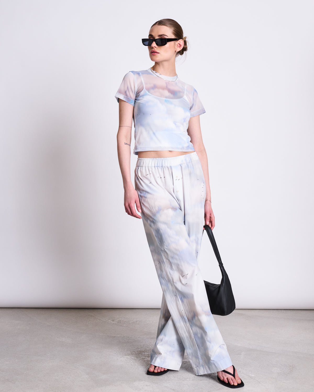 A woman wears the jan-n-june MESH T-SHIRT EVIE CLOUD PRINT with matching wide-leg pants. Perfect for festivals, she pairs her recycled mesh outfit with sunglasses, black sandals, and a black bag, standing confidently against a plain background.