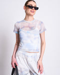 A woman in jan-n-june’s MESH T-SHIRT EVIE CLOUD PRINT over a white top, loose matching pants, black sunglasses, and holding a black bag smiles against a plain white background.