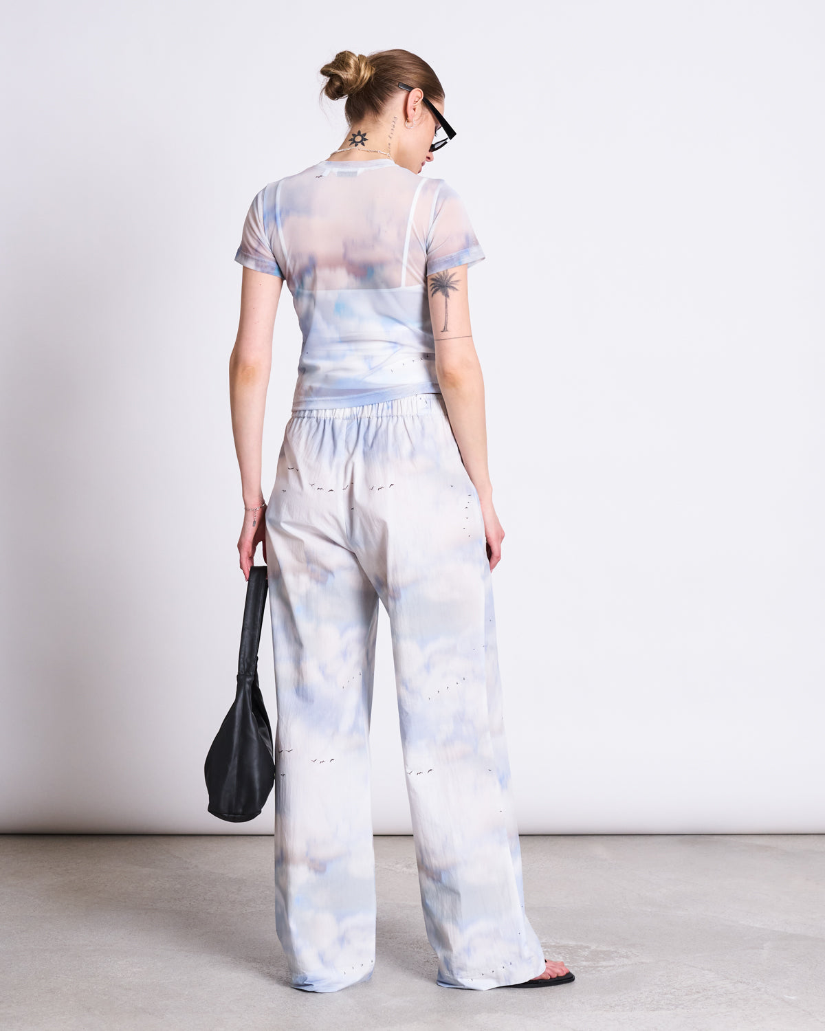 A woman with a blonde bun, sunglasses, and visible tattoos stands with her back to the camera wearing the jan-n-june MESH T-SHIRT EVIE CLOUD PRINT, styled as sheer tie-dye festival wear, holding a black bag against a white background.