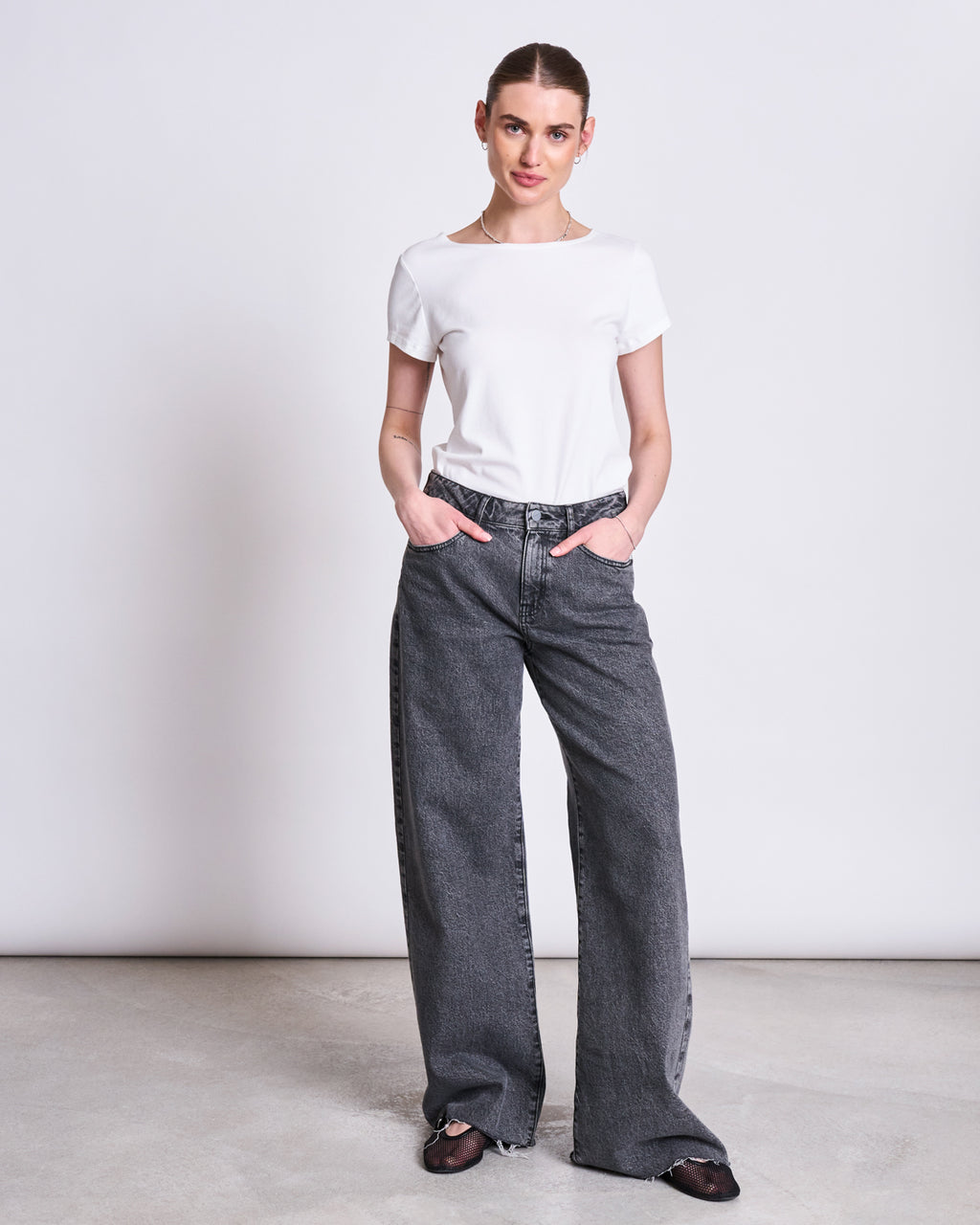 A woman stands against a plain background wearing the jan-n-june T-SHIRT OTTO WHITE GOTS, high-waisted loose gray jeans, and black shoes. With her hands in her pockets, she looks at the camera with a slight smile.