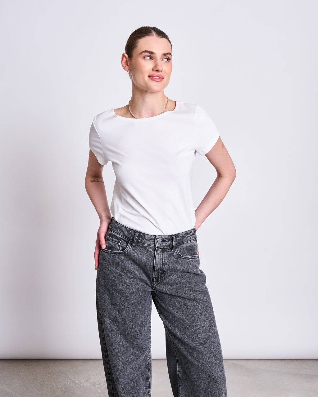 A woman stands against a plain background wearing the jan-n-june T-SHIRT OTTO WHITE GOTS and high-waisted, loose gray jeans. She looks to the side with her hands on her hips, hair pulled back.