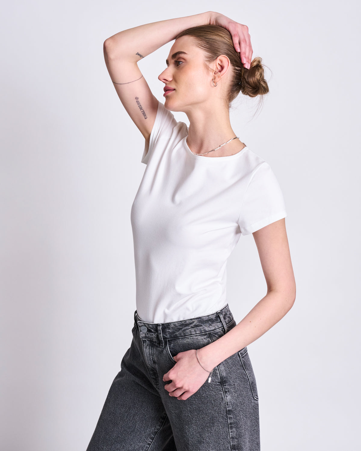 A woman with light skin and a bun poses against a plain background, wearing the jan-n-june T-SHIRT OTTO WHITE GOTS and high-waisted gray jeans, with one arm raised and bent over her head.