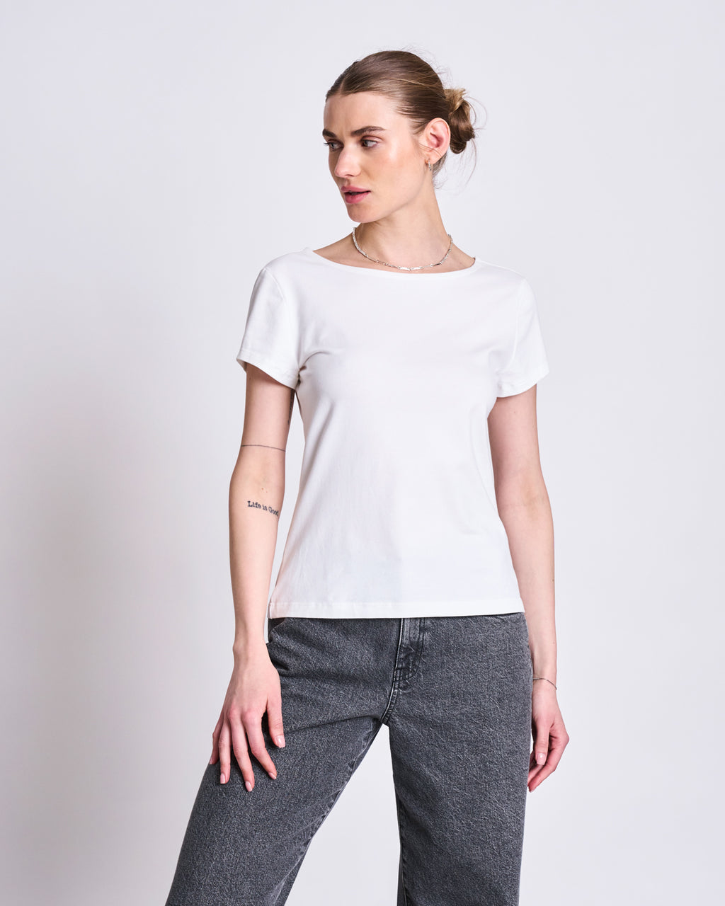 A woman with light skin and brown hair in a bun wears the jan-n-june T-SHIRT OTTO WHITE GOTS, paired with gray jeans, standing against a plain light background and looking to her left.