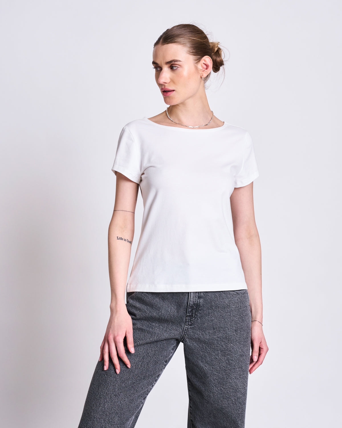 A woman with light skin and brown hair in a bun wears the jan-n-june T-SHIRT OTTO WHITE GOTS, paired with gray jeans, standing against a plain light background and looking to her left.