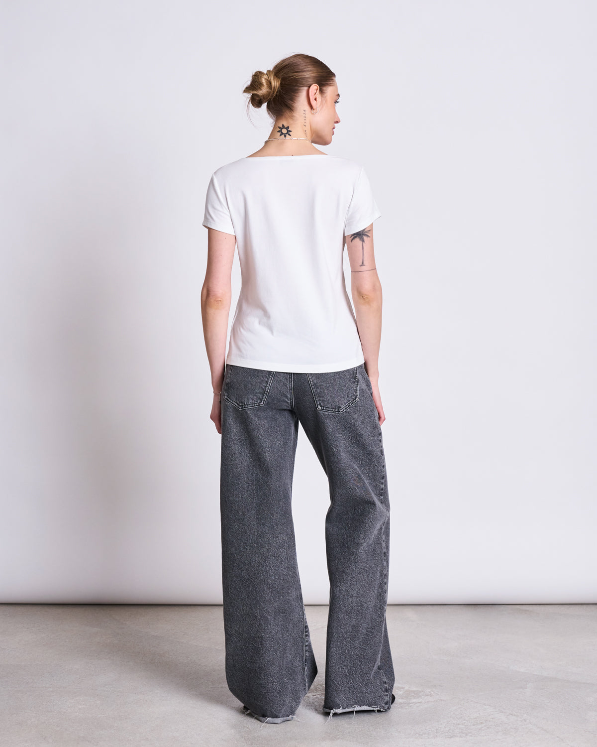 A person with a tattooed neck and arm faces away, wearing the jan-n-june T-SHIRT OTTO WHITE GOTS and loose, dark gray jeans with frayed hems against a plain background.