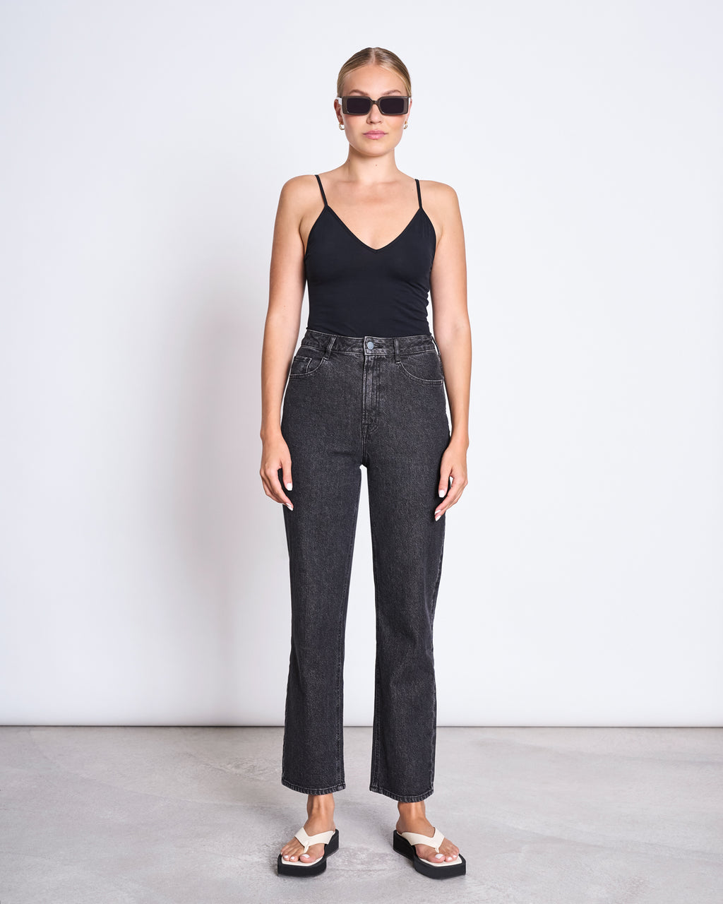 A woman stands against a white background wearing sunglasses and the jan-n-june TOP MATILDA BLACK GOTS, paired with high-waisted dark jeans and black-and-white sandals. Her hair is tied back, and her arms are relaxed at her sides.