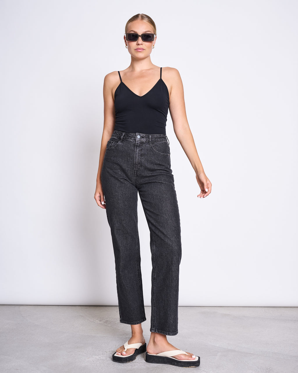 A woman wearing dark sunglasses, high-waisted black jeans, white sandals, and the jan-n-june TOP MATILDA BLACK GOTS—a V-neck sleeveless top made from organic cotton—stands against a plain white background.