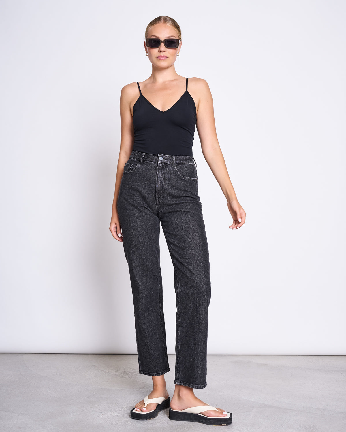 A woman wearing dark sunglasses, high-waisted black jeans, white sandals, and the jan-n-june TOP MATILDA BLACK GOTS—a V-neck sleeveless top made from organic cotton—stands against a plain white background.