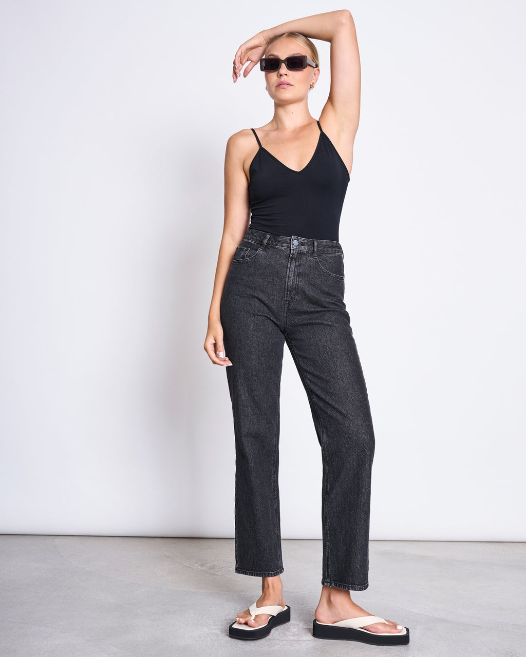 A woman in sunglasses, the jan-n-june TOP MATILDA BLACK GOTS sleeveless top, high-waisted black jeans, and white sandals stands against a white wall with one arm raised and resting on her head.