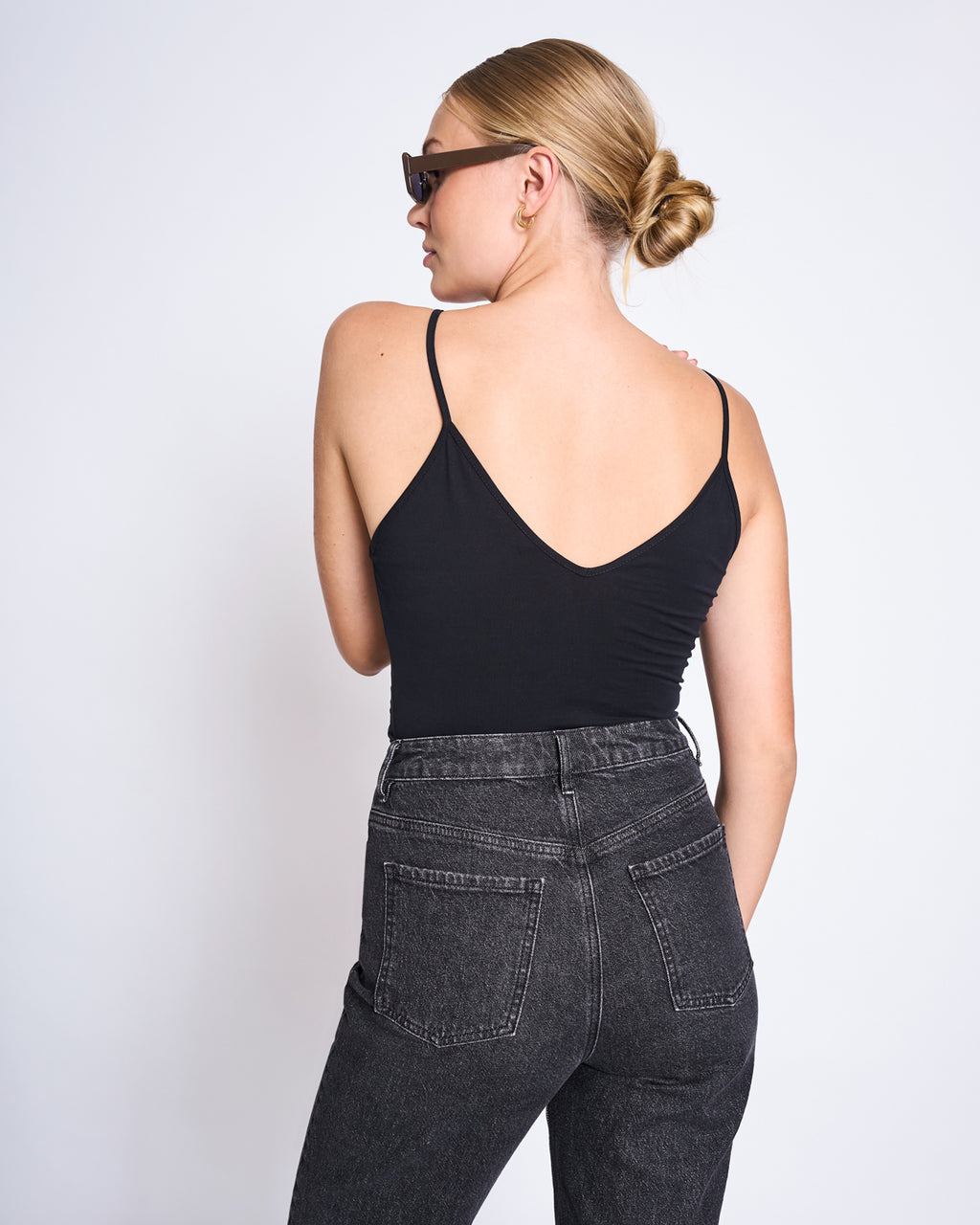 A woman with blonde hair in a bun, dark sunglasses, and high-waisted black jeans stands with her back to the camera wearing the jan-n-june TOP MATILDA BLACK GOTS against a plain white background.