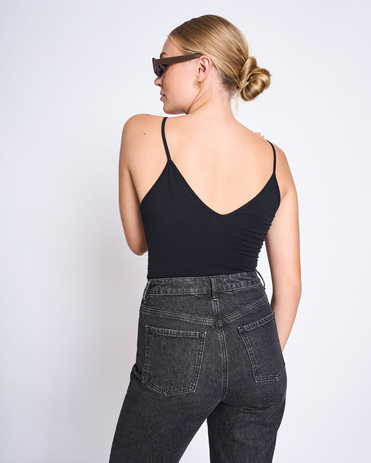 A woman with blonde hair in a bun, dark sunglasses, and high-waisted black jeans stands with her back to the camera wearing the jan-n-june TOP MATILDA BLACK GOTS against a plain white background.