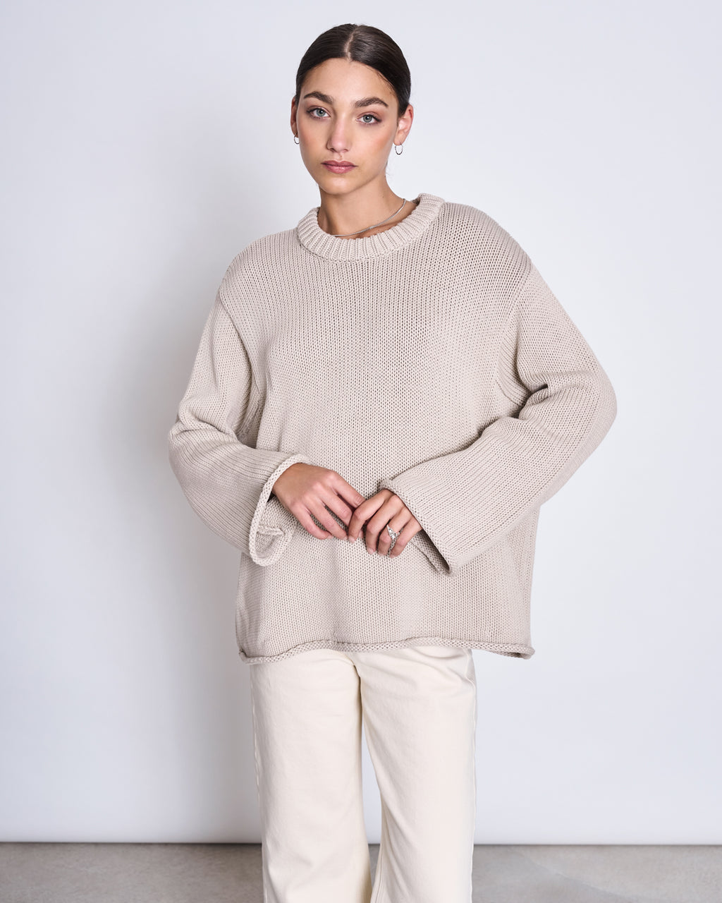 A woman stands against a plain background wearing the jan-n-june JUMPER SONA IVORY GOTS, an oversized knit sweater made from soft, organic cotton, paired with light-colored pants. She has her hands together and gazes to the side neutrally.