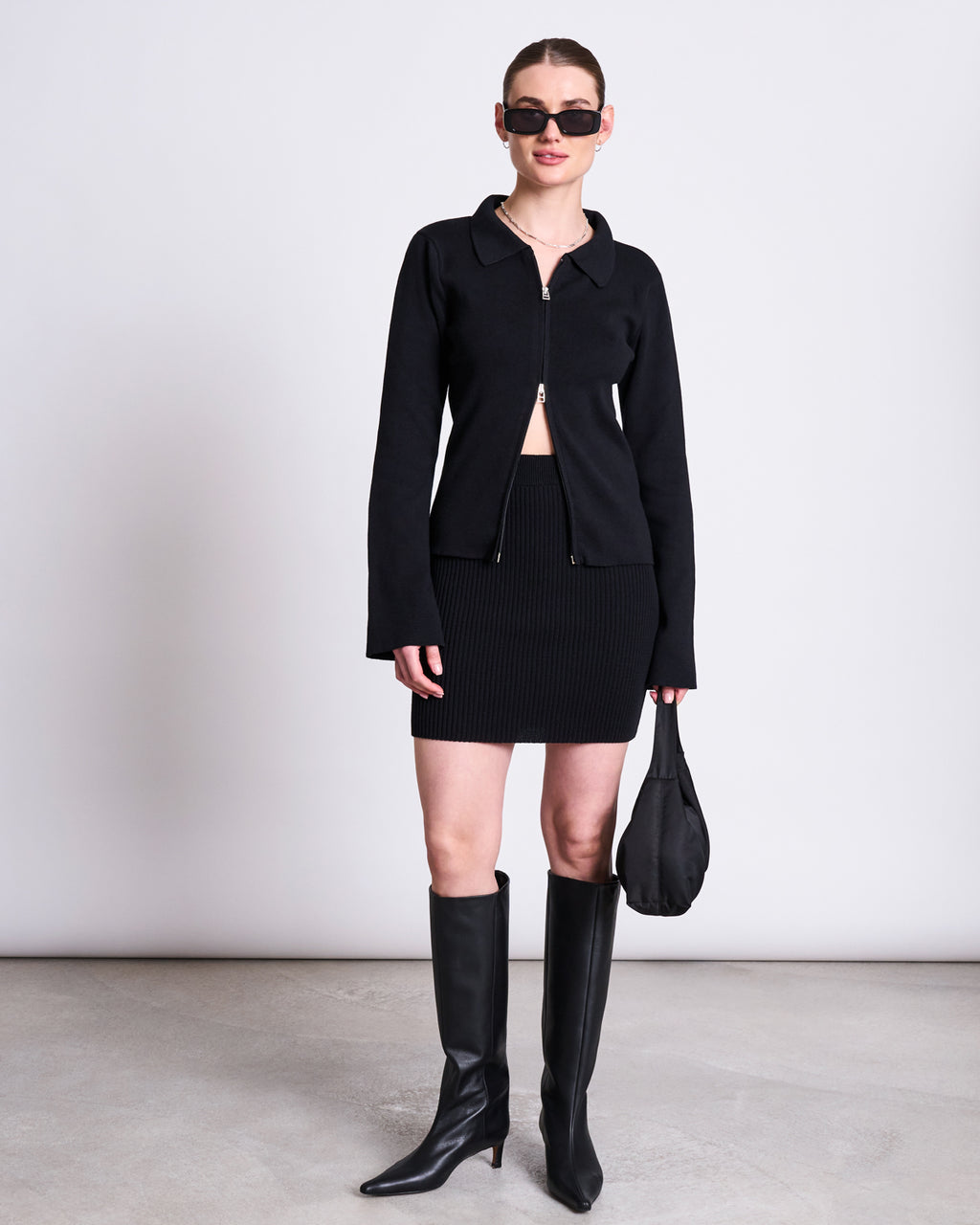 A woman stands against a plain background wearing jan-n-june’s SKIRT LOKKA BLACK GOTS with an elasticated waistband, a black zip-up jacket, tall black boots, sunglasses, and holding a small black handbag.