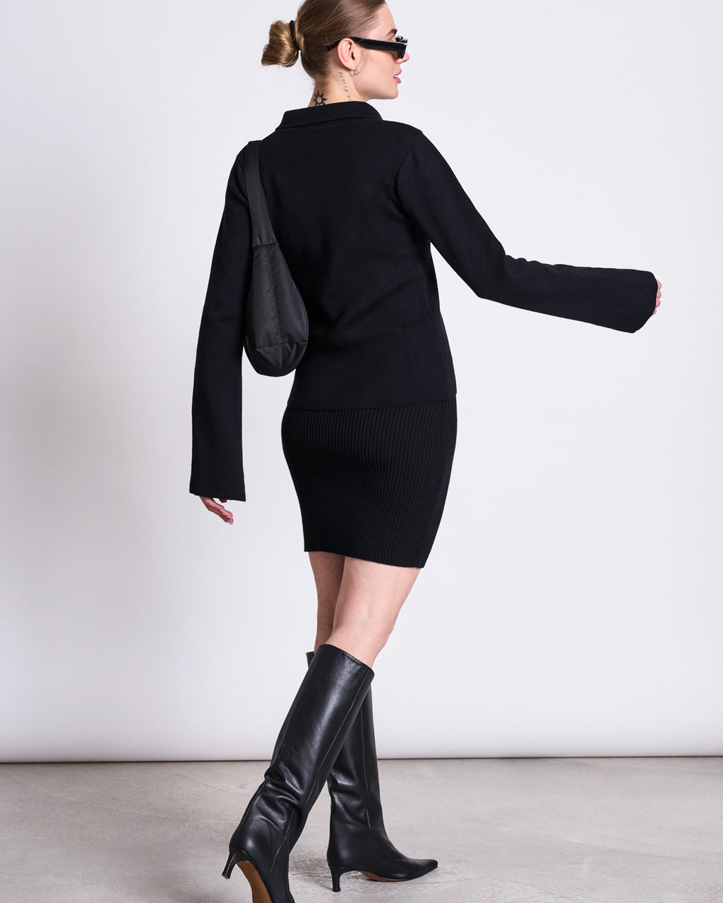 A woman in a black long-sleeve top, jan-n-june’s SKIRT LOKKA BLACK GOTS with an elastic waistband, black knee-high boots, sunglasses, and a black shoulder bag walks away from the camera against a plain background.