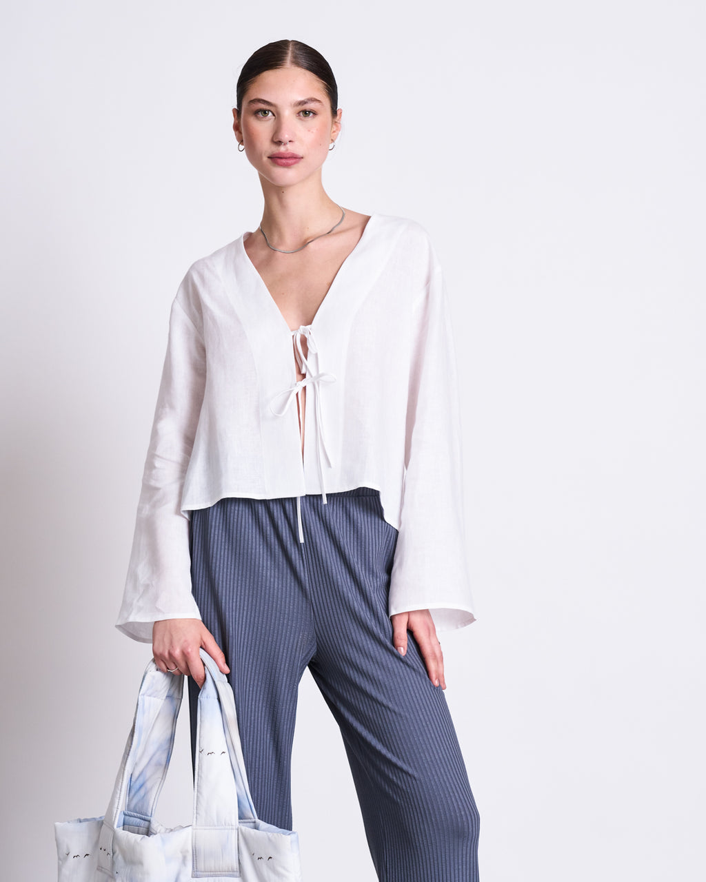 A woman stands against a plain background wearing the jan-n-june LINEN TIE BLOUSE SKAGEN WHITE with blue pinstriped pants, holding a light-colored tote bag in her left hand.