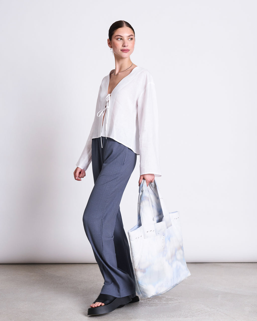 A woman stands against a plain background wearing the jan-n-june LINEN TIE BLOUSE SKAGEN WHITE, paired with dark gray pants, black open-toe sandals, and holding a large white tote bag with a blue tie-dye pattern.