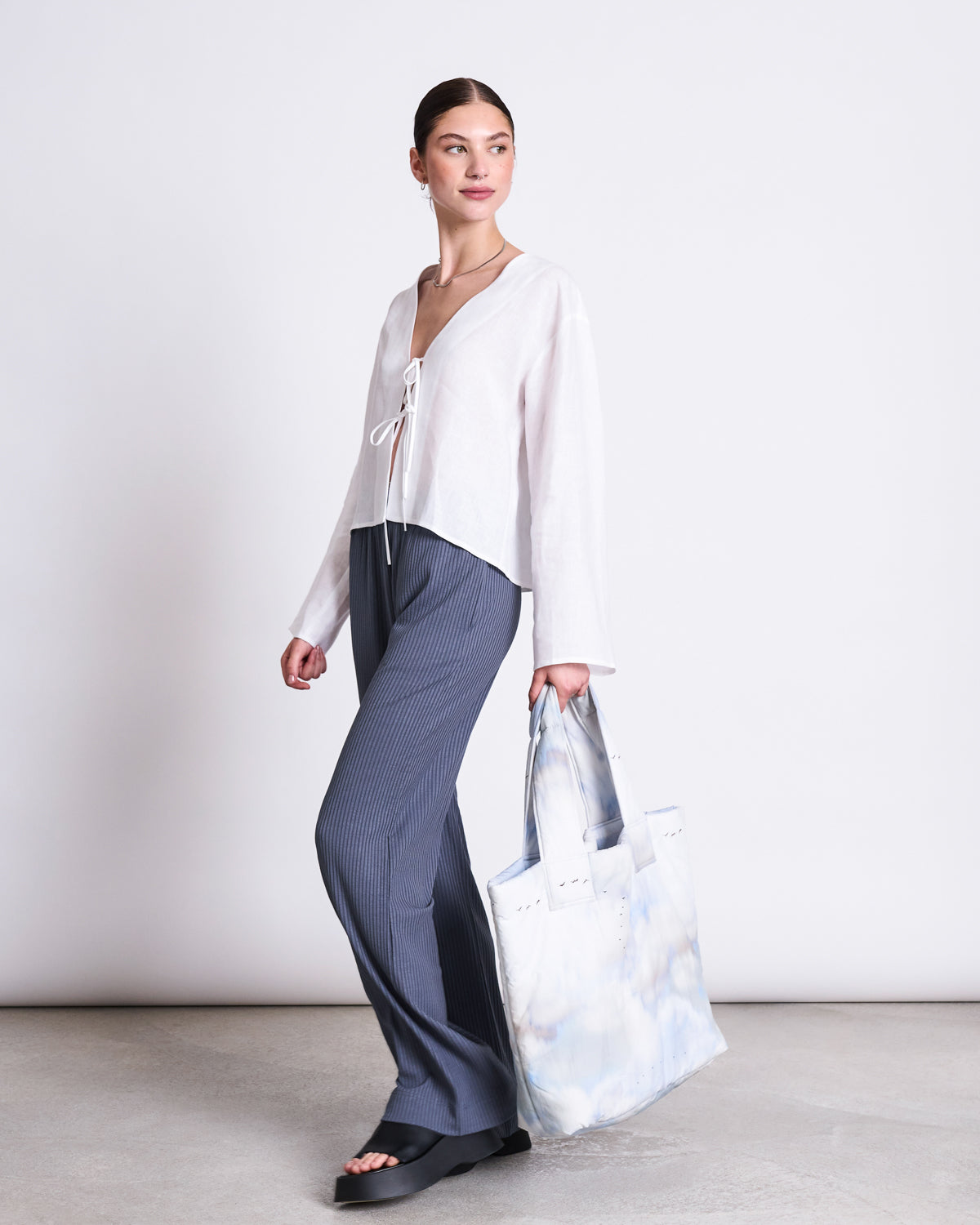 A woman stands against a plain background wearing the jan-n-june LINEN TIE BLOUSE SKAGEN WHITE, paired with dark gray pants, black open-toe sandals, and holding a large white tote bag with a blue tie-dye pattern.
