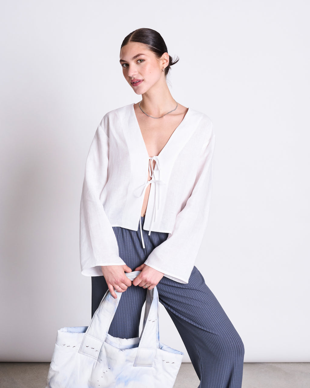 A woman with dark hair in a low bun wears a white, long-sleeve, tie-front blouse and jan-n-june’s LYOCELL RIB PANTS SMILLA TURBULENCE. She holds a large light tote and stands against a plain white background, smiling softly.