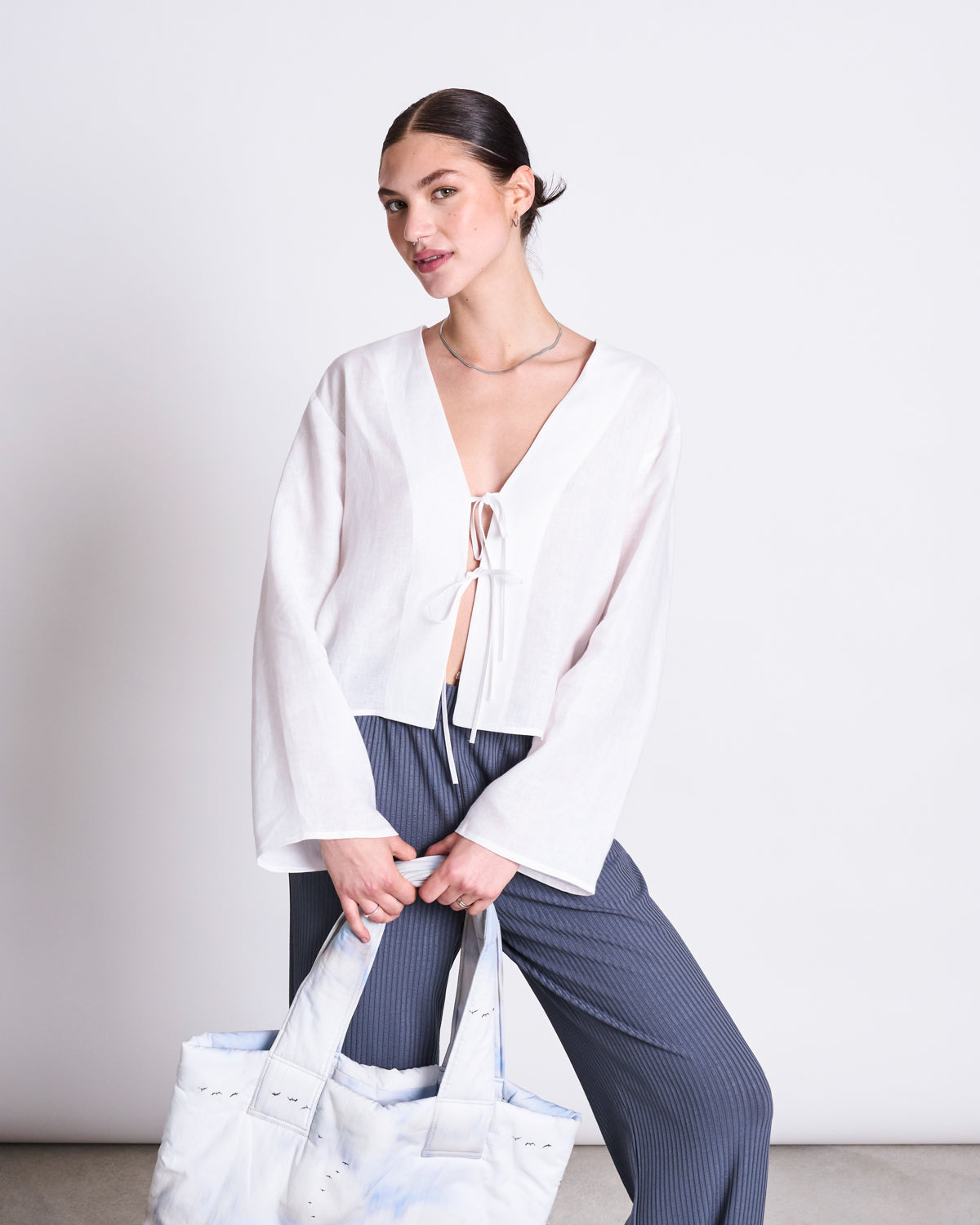 A woman with dark hair in a low bun wears a white, long-sleeve, tie-front blouse and jan-n-june’s LYOCELL RIB PANTS SMILLA TURBULENCE. She holds a large light tote and stands against a plain white background, smiling softly.