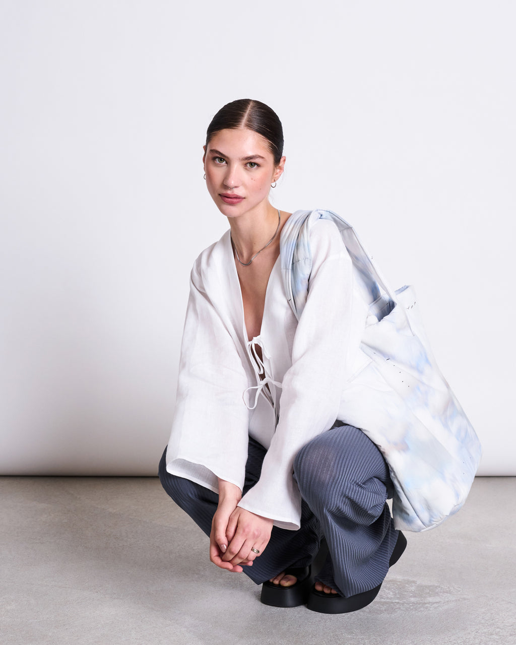 A woman with dark hair slicked back squats on the floor, wearing a white blouse, jan-n-june LYOCELL RIB PANTS SMILLA TURBULENCE, and black sandals. She carries a large tie-dye tote bag over one shoulder, set against a plain white background.
