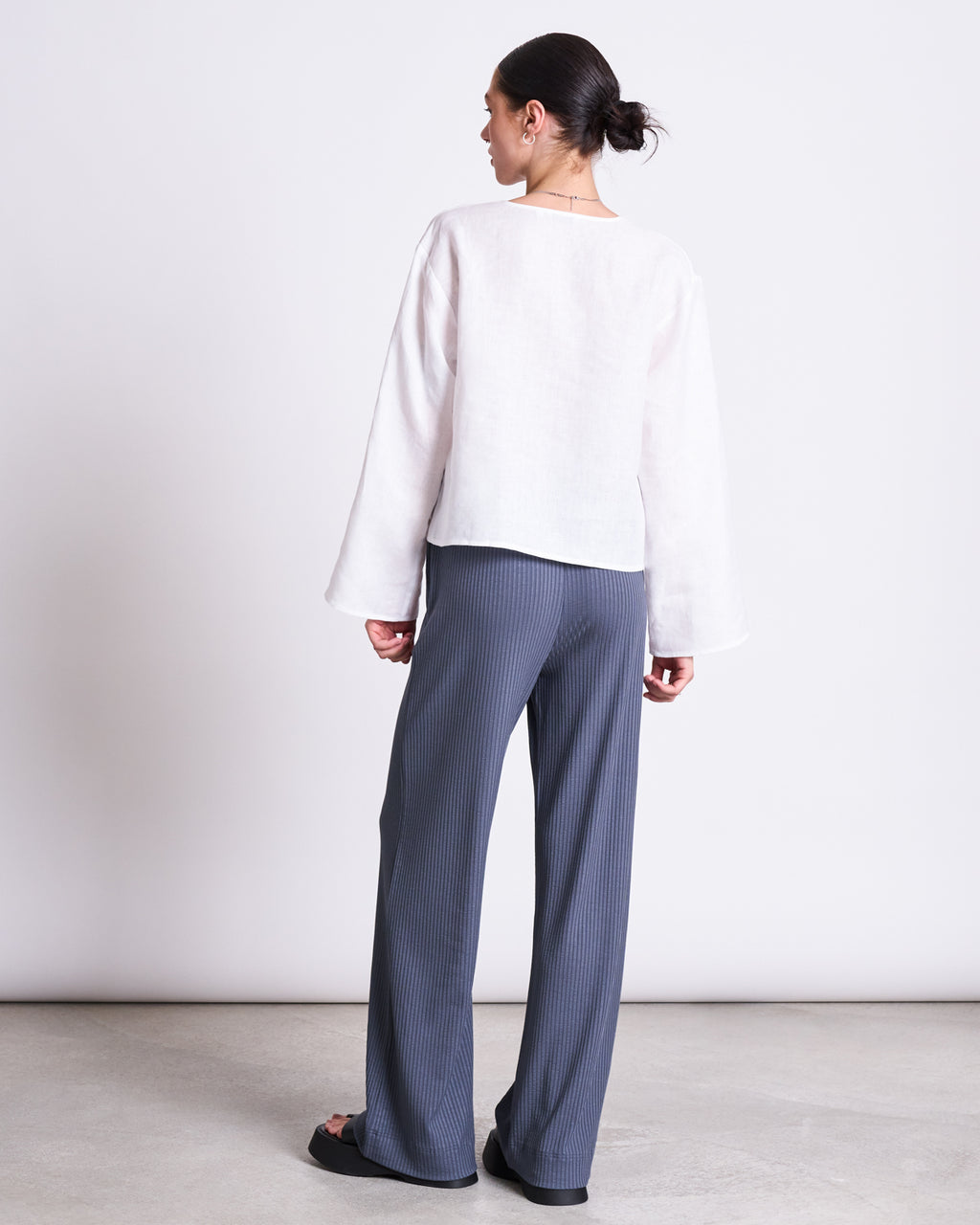 A person faces away from the camera, wearing the jan-n-june LINEN TIE BLOUSE SKAGEN WHITE and gray wide-leg pants with subtle vertical stripes, set against a plain light background.
