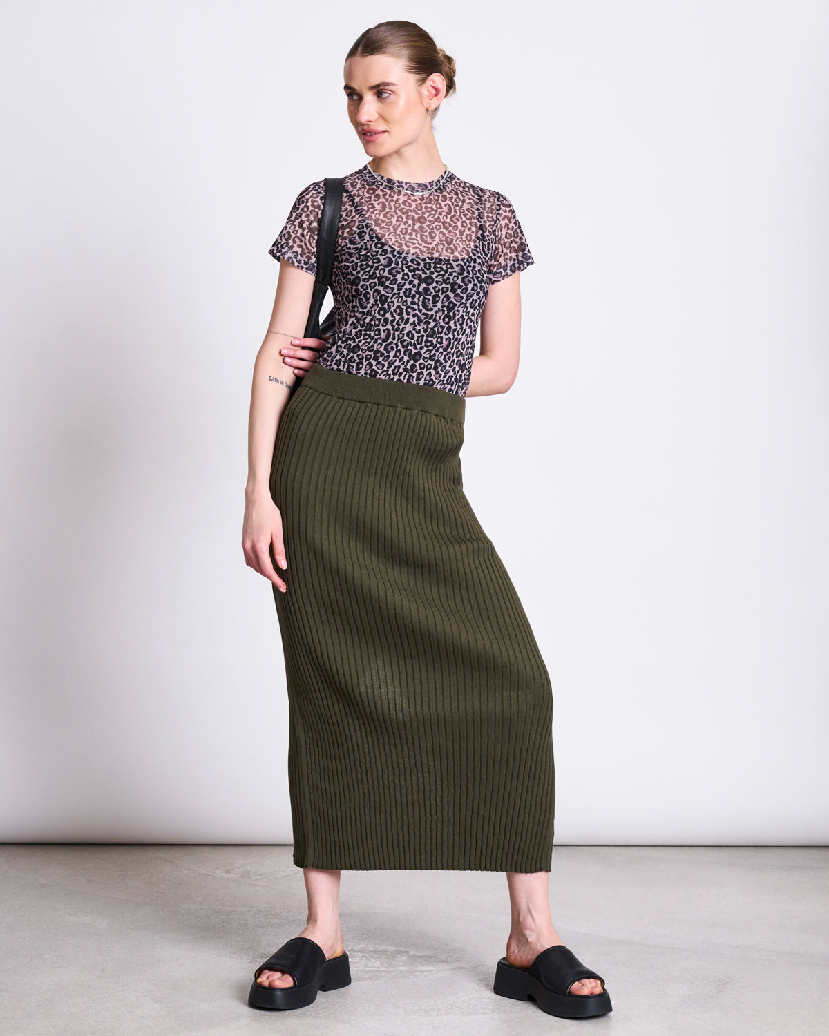 A woman with her hair tied back wears the jan-n-june MESH T-SHIRT EVIE LEO PRINT over a black tank, styled with an olive ribbed midi skirt, black slides, and a black shoulder bag—ideal for bold festival looks.