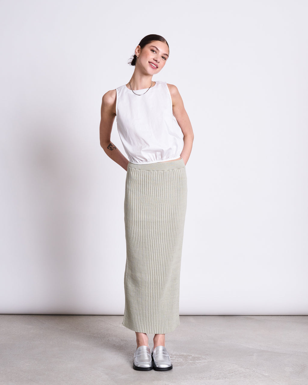 A woman poses against a white background, wearing jan-n-junes MIDI KNIT SKIRT EBBA VINTAGE LACE GOTS—a high-waisted, light gray ribbed skirt—paired with a sleeveless white top and silver shoes, her hands in her pockets and smiling slightly.