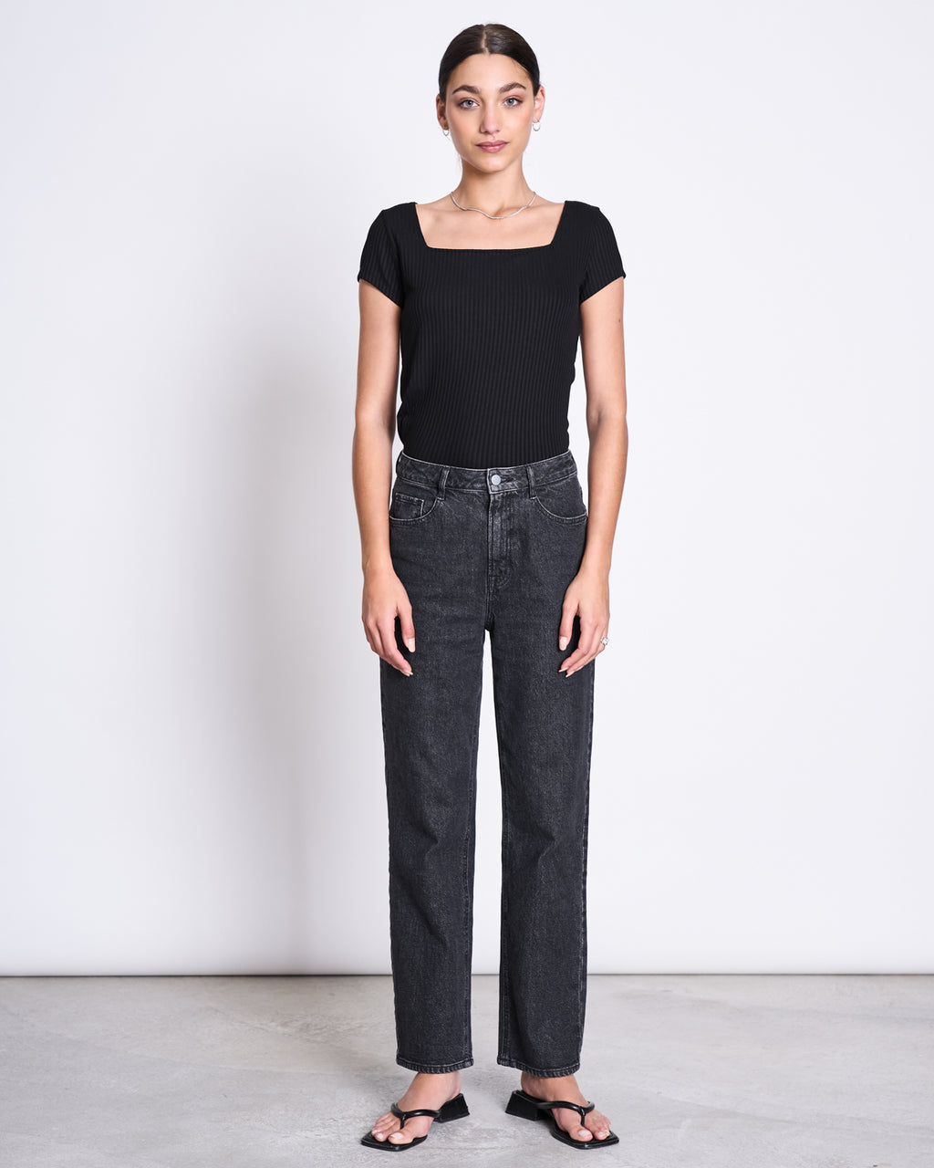 A woman stands against a white background, wearing the jan-n-june T-SHIRT ARIMA BLACK— a fitted, square-neck top in soft ribbed Tencel™. She pairs it with high-waisted black jeans and sandals, facing forward with relaxed arms at her sides.