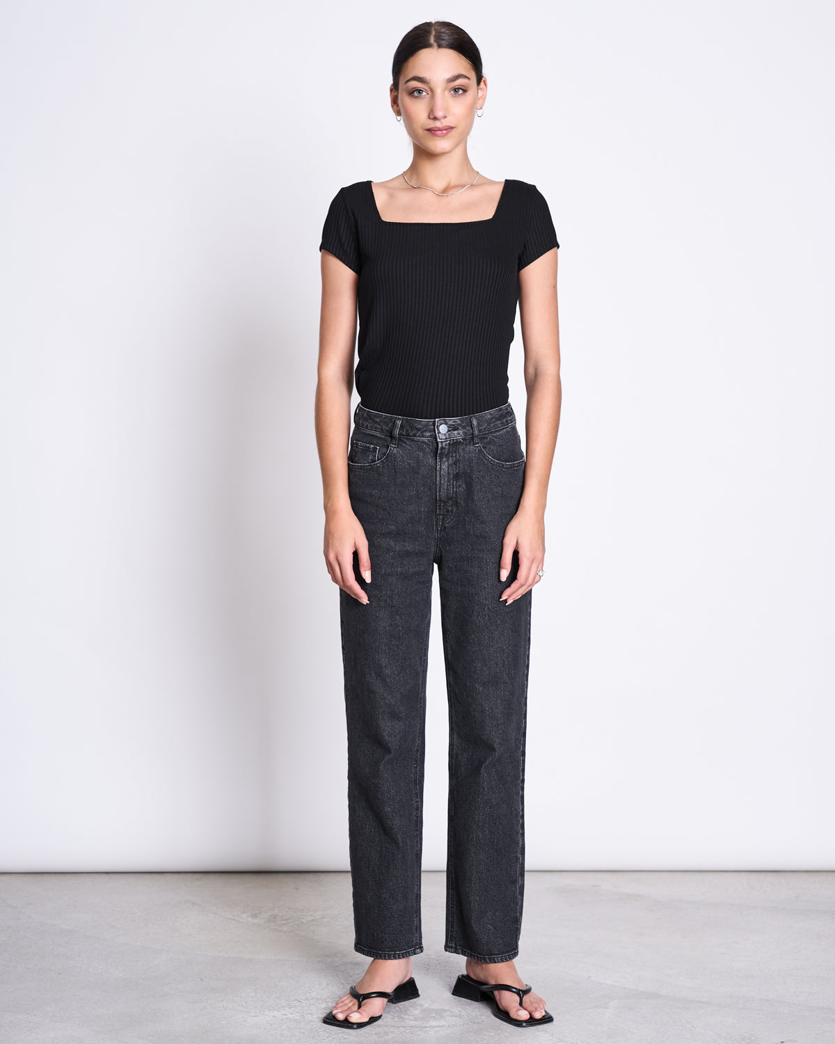 A woman stands against a white background, wearing the jan-n-june T-SHIRT ARIMA BLACK— a fitted, square-neck top in soft ribbed Tencel™. She pairs it with high-waisted black jeans and sandals, facing forward with relaxed arms at her sides.