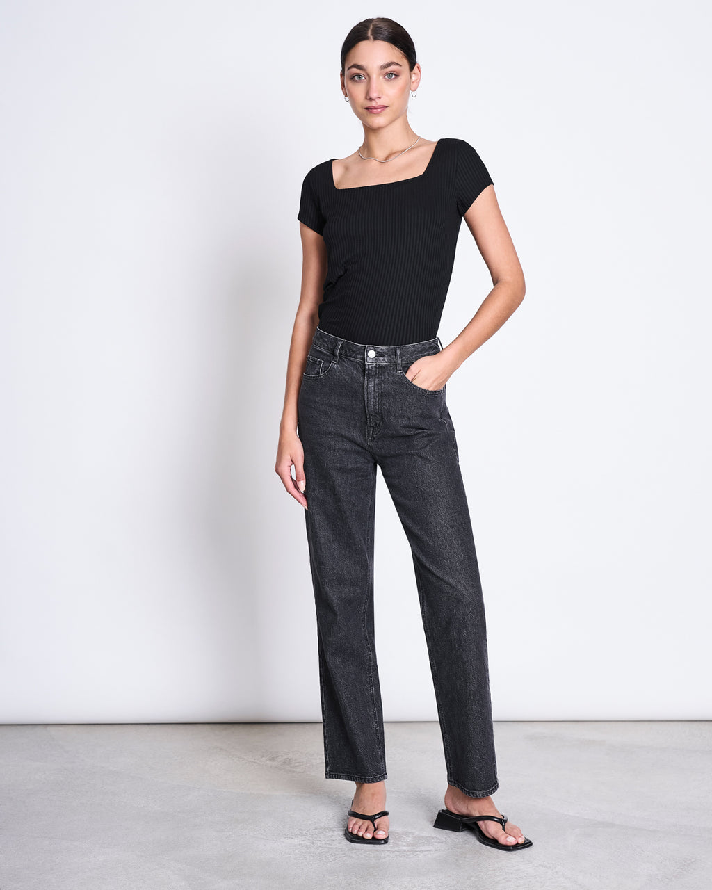 A woman stands against a white wall wearing the jan-n-june T-SHIRT ARIMA BLACK, styled with high-waisted straight-leg black jeans and black sandals. One hand is in her pocket and she has a neutral expression.