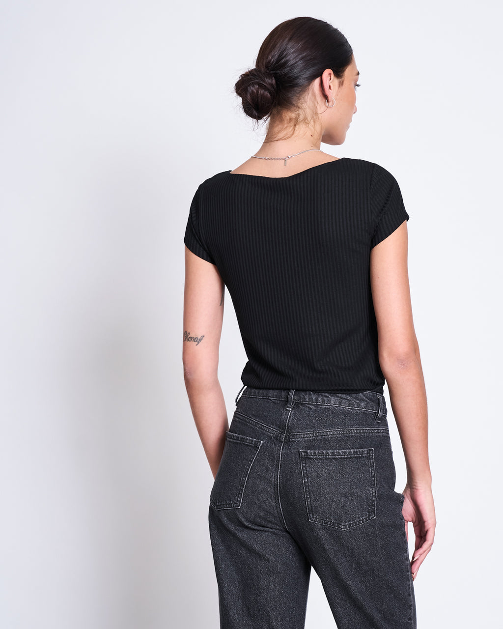 A woman with dark hair in a low bun is seen from the back, wearing the jan-n-june T-SHIRT ARIMA BLACK— a short-sleeve, square neck top made from Tencel™ rib fabric—paired with high-waisted dark gray jeans against a white background.