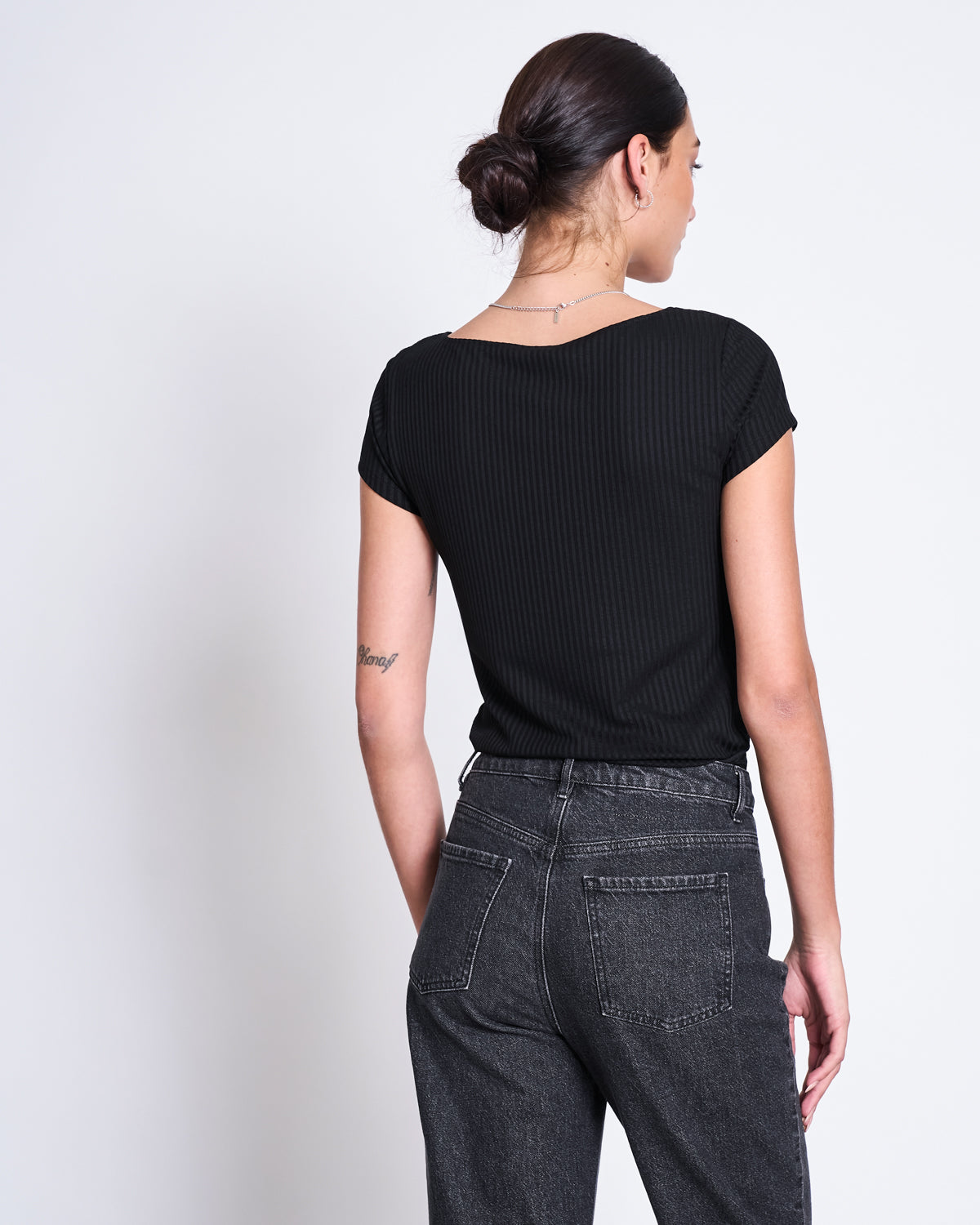 A woman with dark hair in a low bun is seen from the back, wearing the jan-n-june T-SHIRT ARIMA BLACK— a short-sleeve, square neck top made from Tencel™ rib fabric—paired with high-waisted dark gray jeans against a white background.