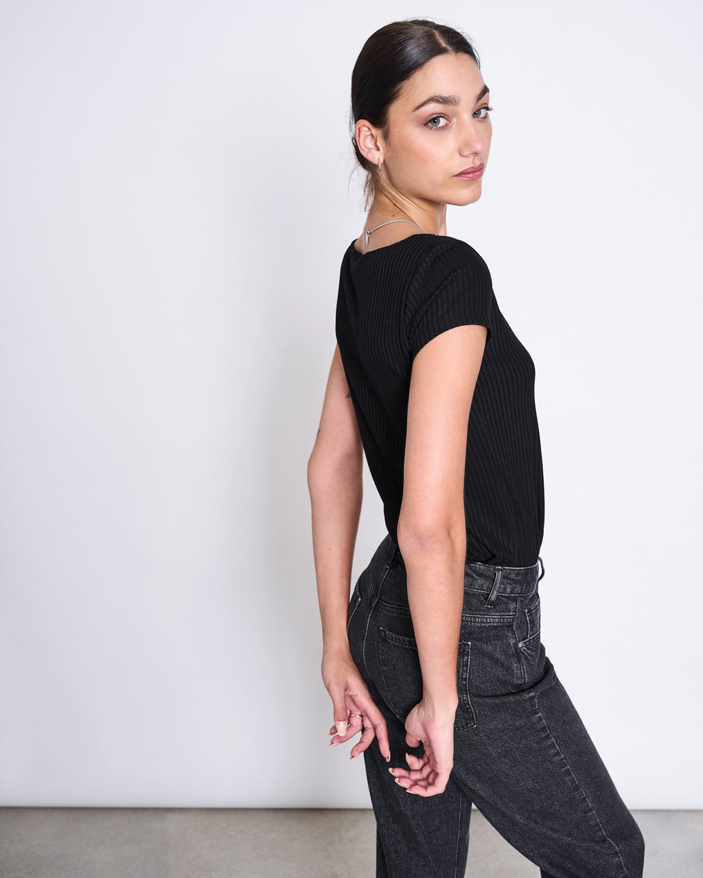 A woman with dark hair tied back models the jan-n-june T-SHIRT ARIMA BLACK in soft Tencel™ rib fabric, paired with dark jeans. She stands sideways, looking over her shoulder against a plain light background.